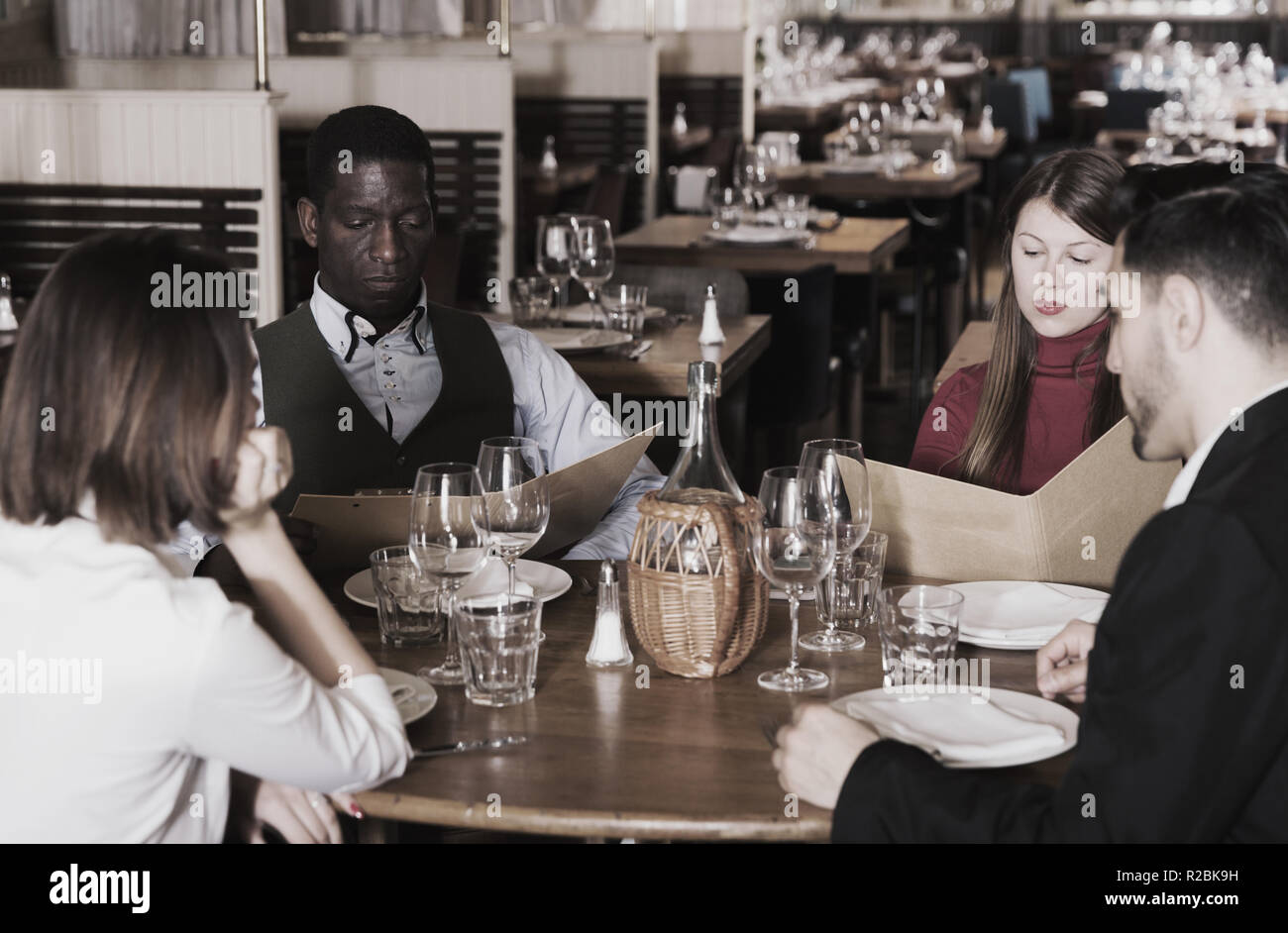 Group of young people sitting at table in cozy restaurant, choosing dishes from menu Stock Photo