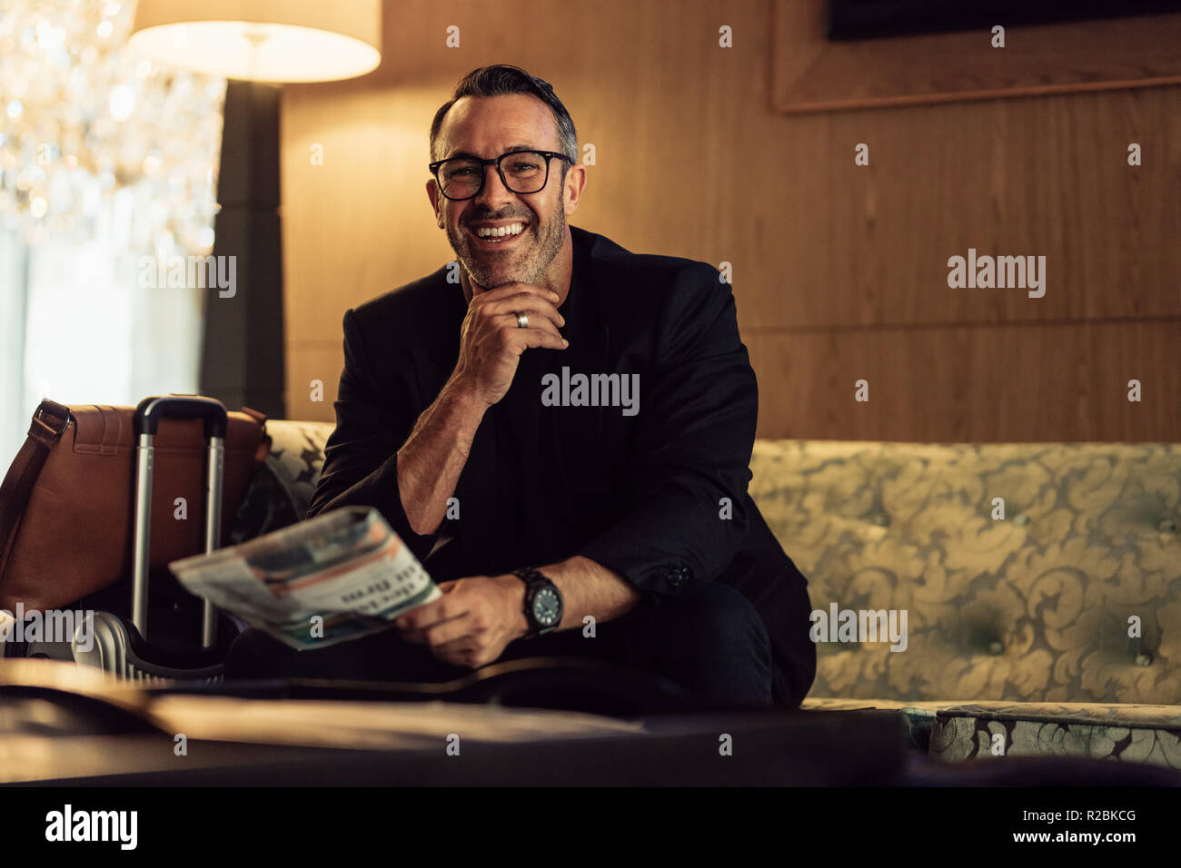 Portrait of happy middle aged businessman sitting in hotel waiting area with his luggage on the side. CEO waiting in hotel lobby. Stock Photo