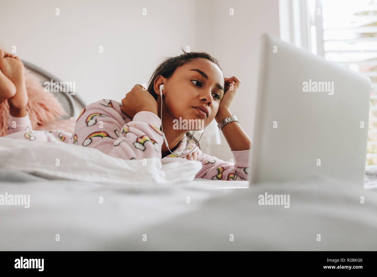 Young girl in bed watching a movie on laptop. Girl using laptop lying on bed while listening to music using earphones. Stock Photo
