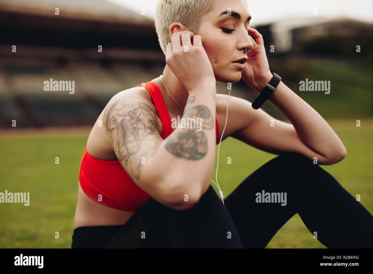 Close up of a woman athlete sitting on ground listening to music wearing earphones. Side view of a fitness woman sitting inside a track and field stad Stock Photo