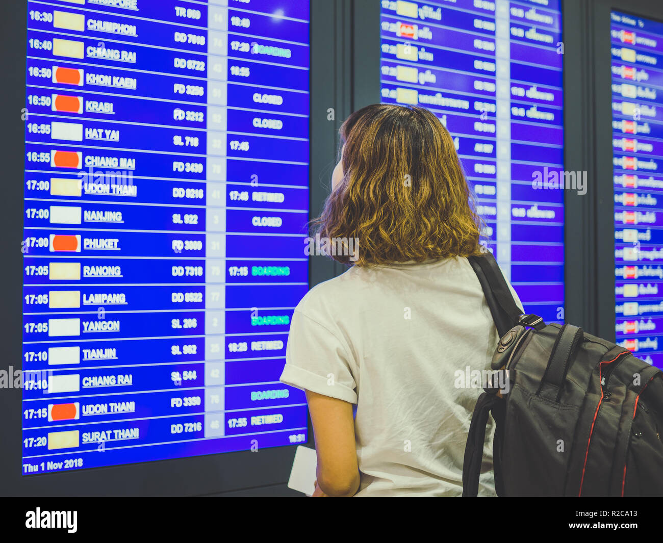 Woman short hair wearing white shirt with backpack checking flight at the flight information board in international airport terminal. Stock Photo