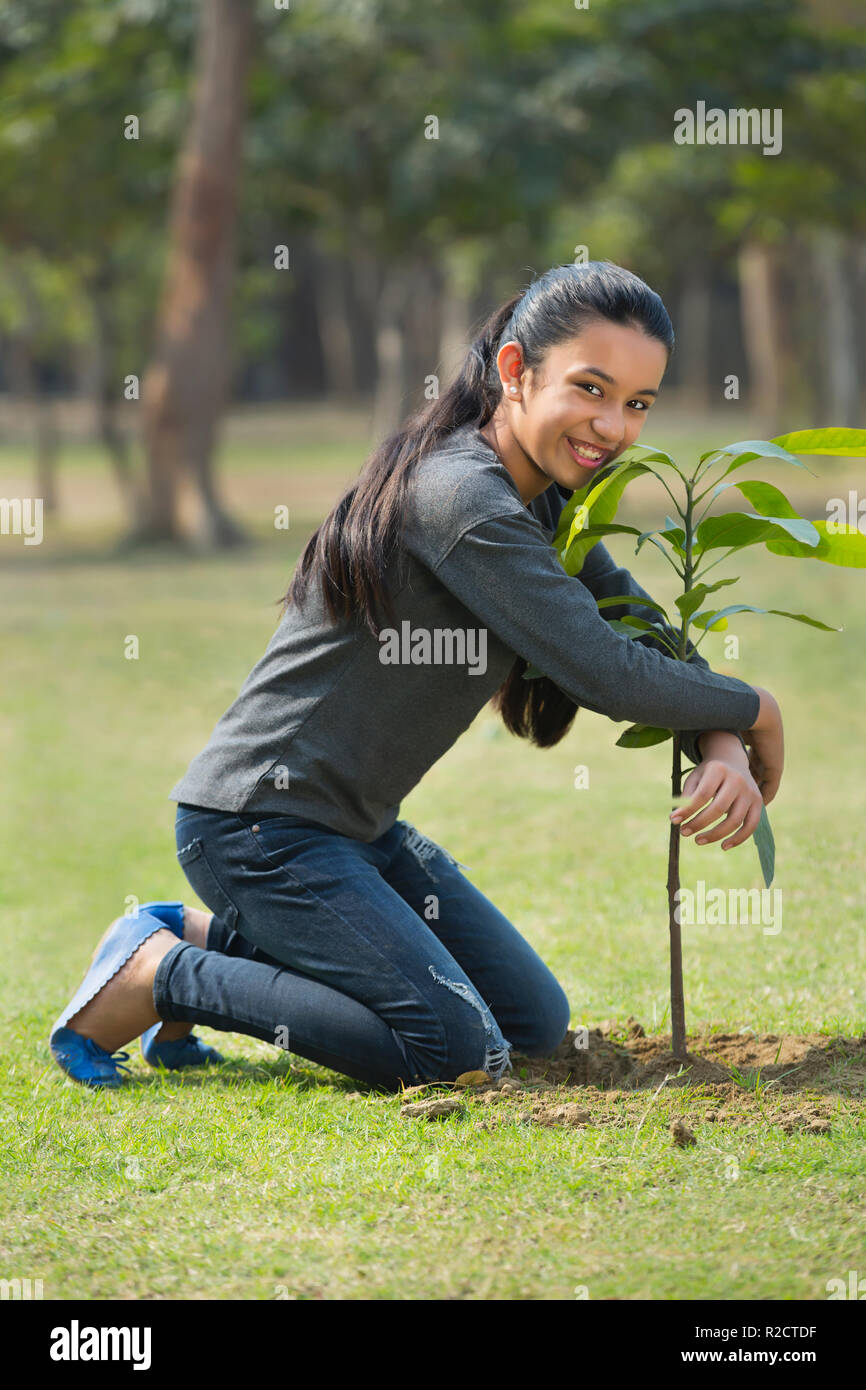 Happy young girl sitting on knees with her arms around a plant in park. Stock Photo