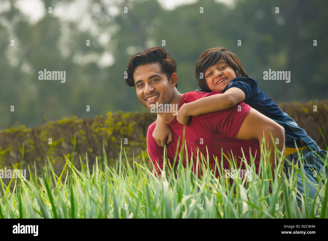 Happy young boy piggy riding on the back of his father outdoors with fresh grass in the foreground. Stock Photo