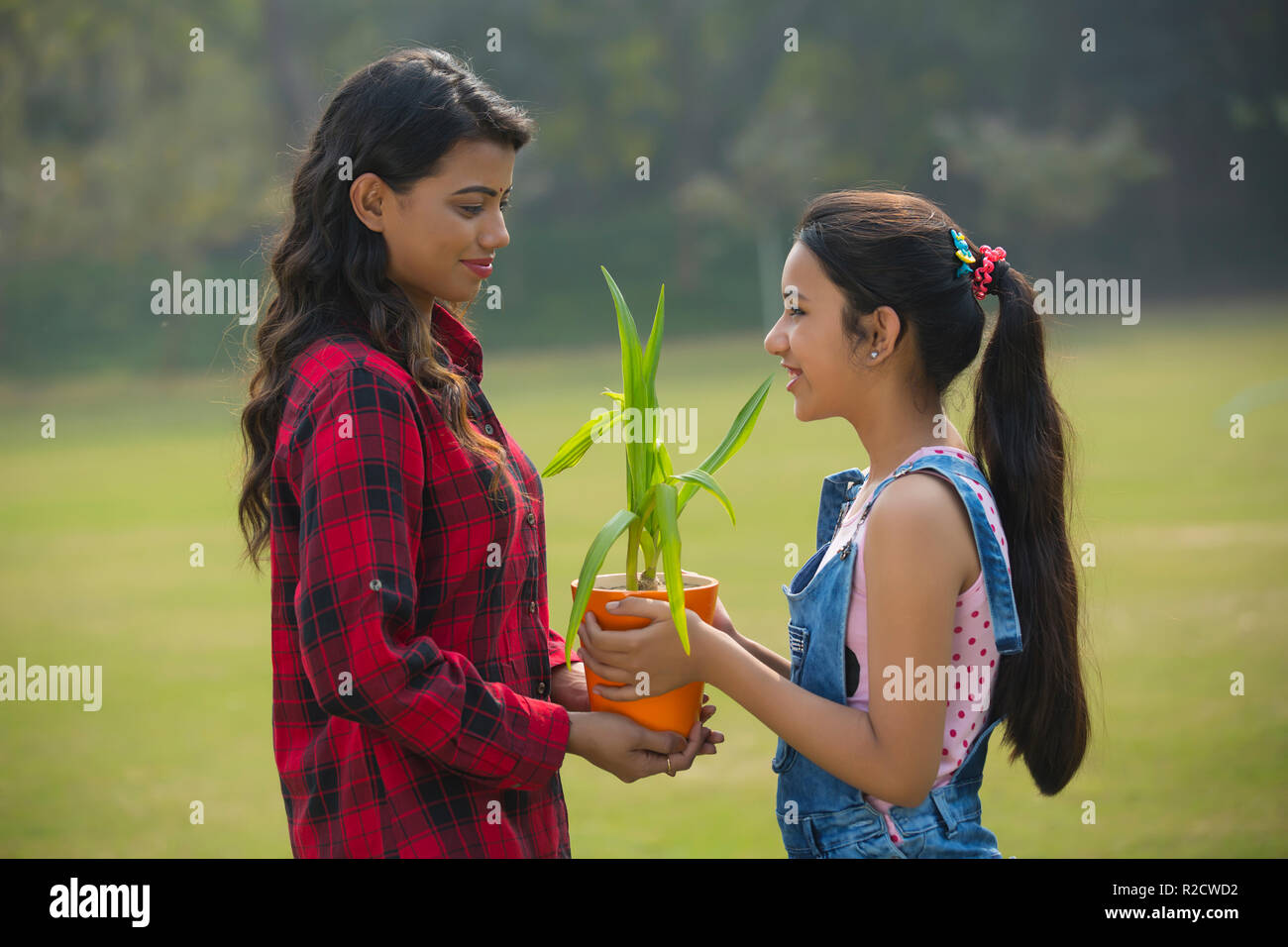 Smiling young girl giving a plant in a flower pot to her mother outdoors. Stock Photo