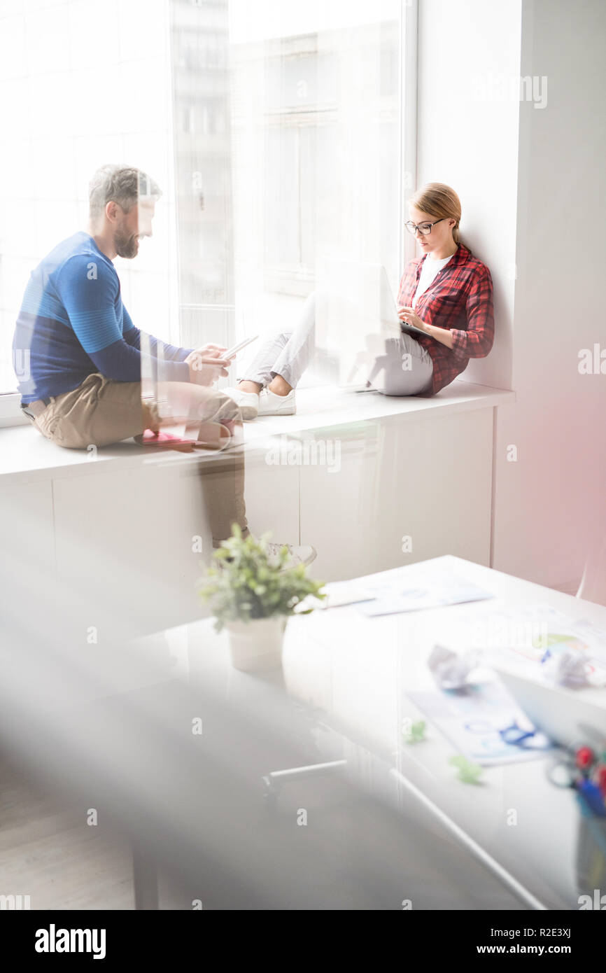 Hipster employees using gadgets while sitting on window sill Stock Photo