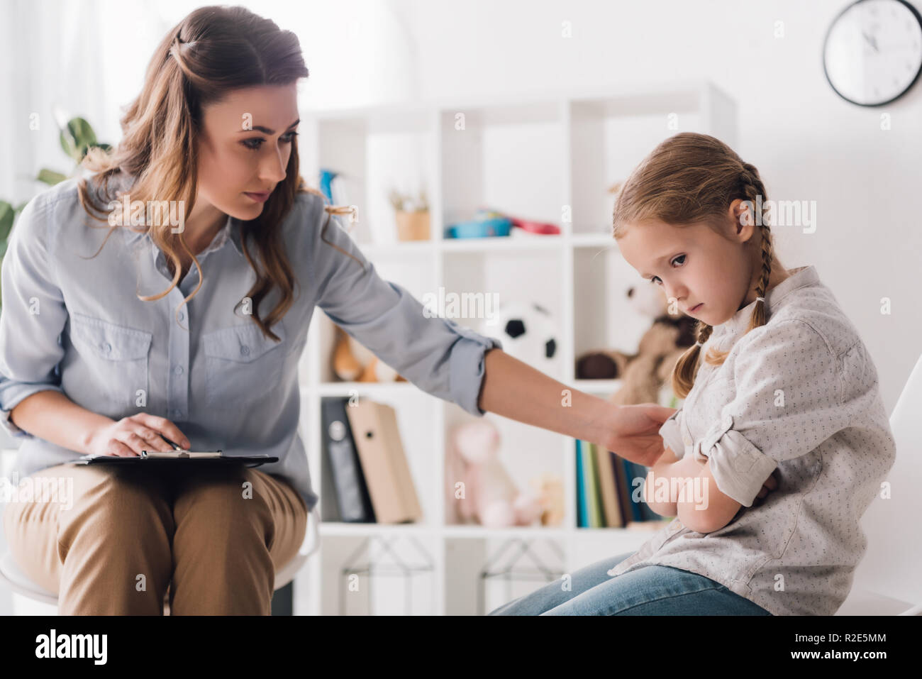 adult psychologist with clipboard talking to sad little child in office Stock Photo
