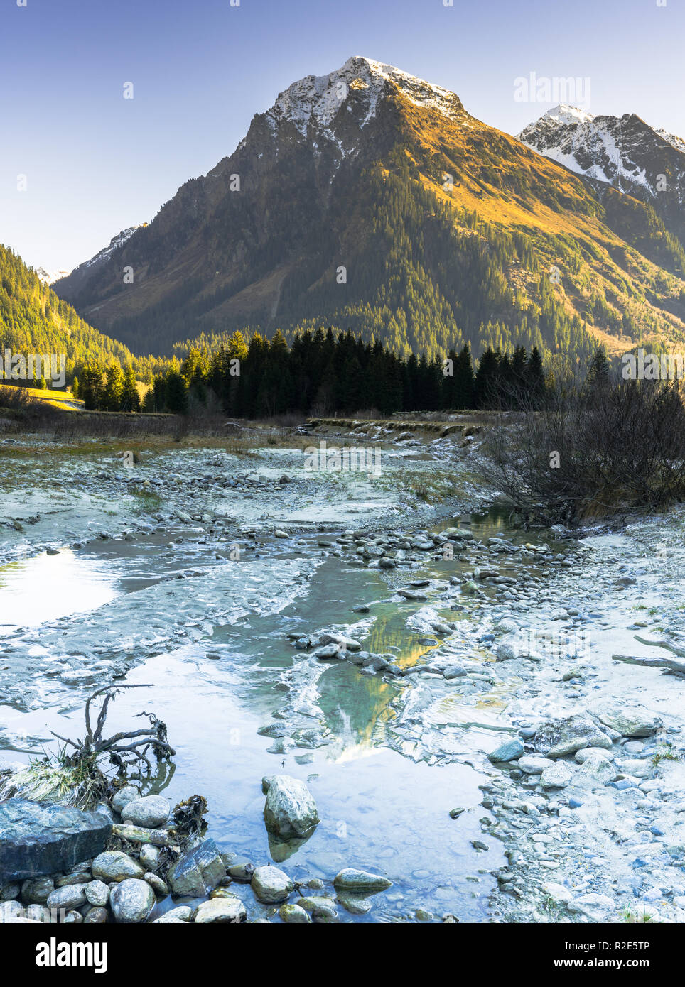 vertical rocky mountain landscape in early winter with snow-capped peaks and colorful forest and a mountain stream in the foreground with ice and rime Stock Photo