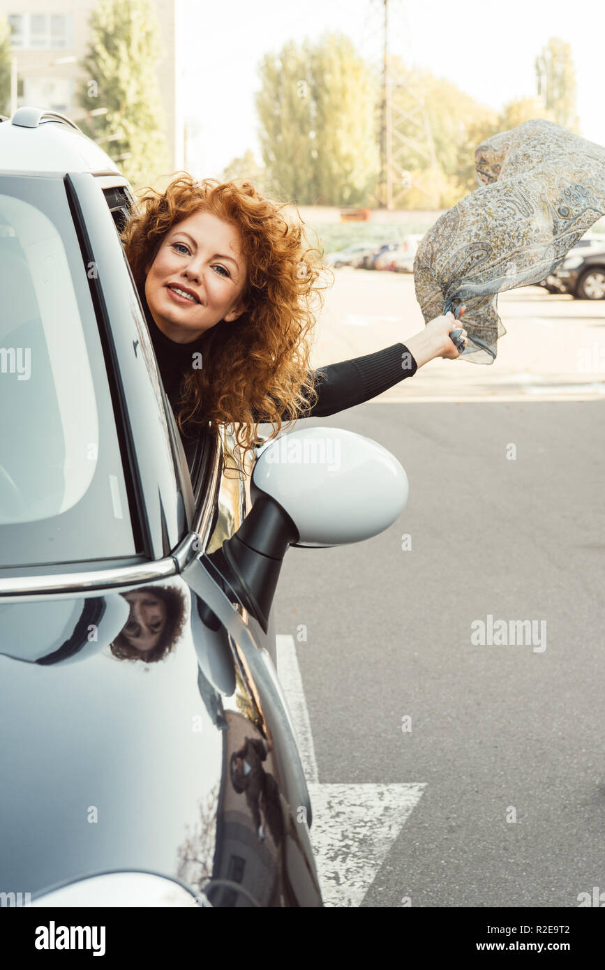 happy curly ginger woman leaning out from car and holding shawl at urban street Stock Photo