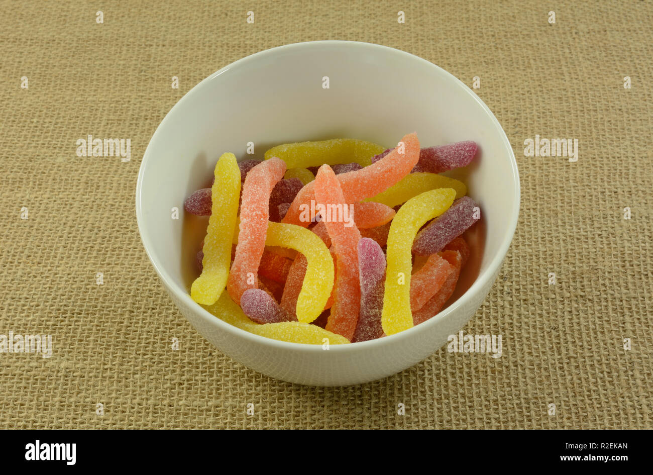 Yellow, orange and purple sour gummy worms candy in white bowl on burlap Stock Photo