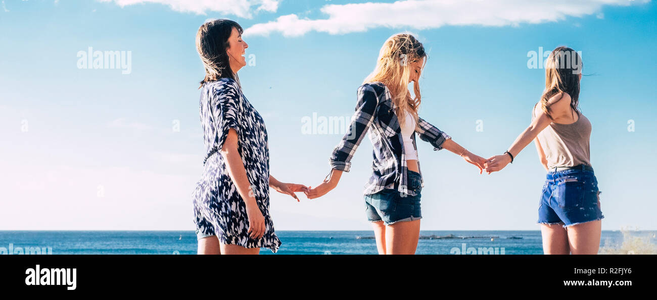 panoramic image with three beautiful young females friends taking hands eachother walking together in friendship with blue sky and ocean in background divided by horizon. Happy people enjoying nature and outdoor leisure activity Stock Photo