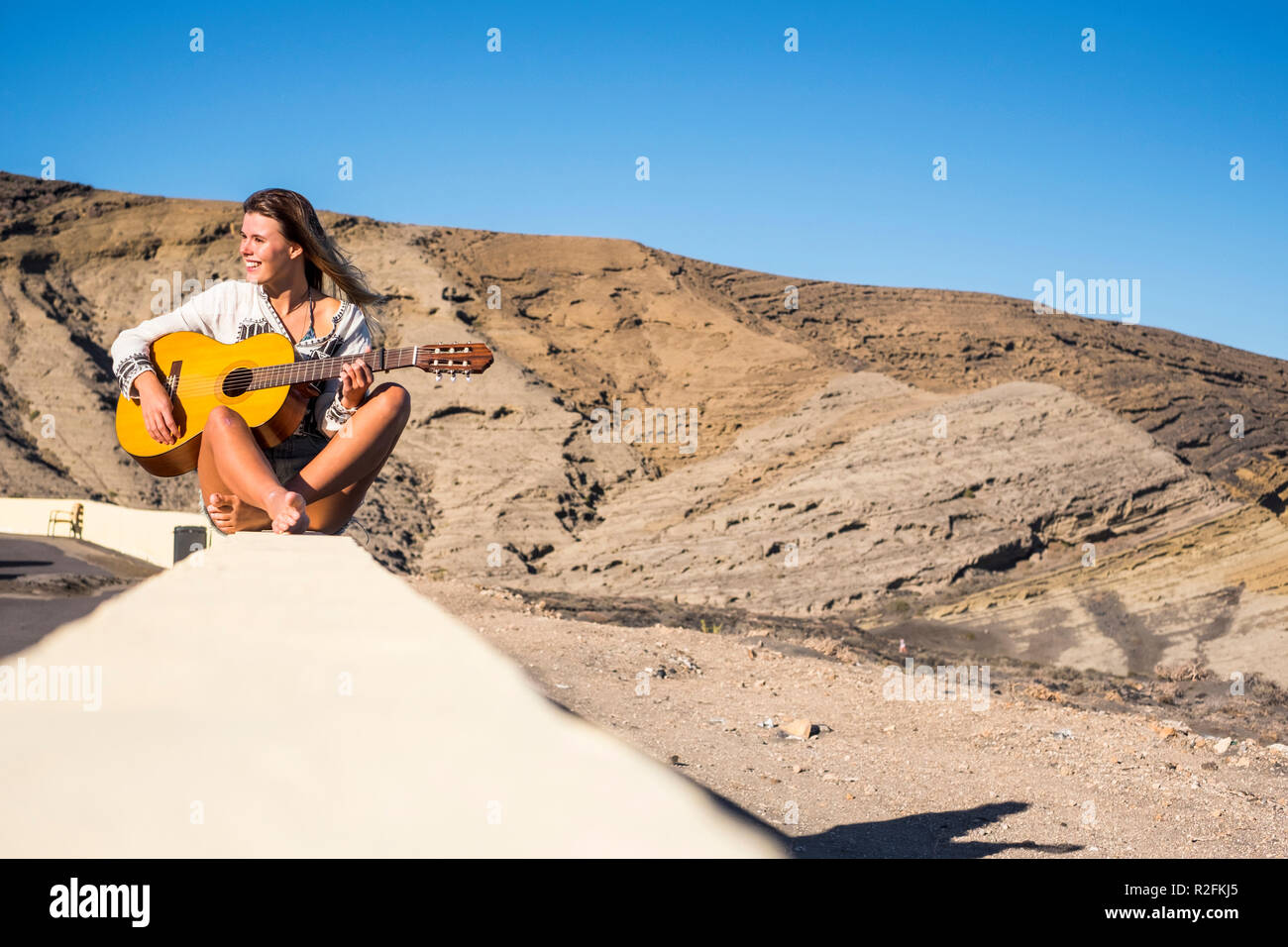 nippy and alternative lifestyle concept for blonde caucasian model smiling outdoor with mountains on the background and playing an old acoustic guitar. wind in her hair. Stock Photo