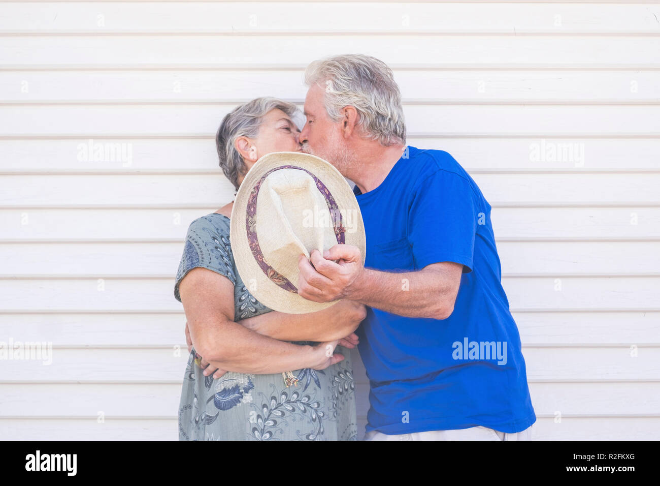 Valentine's day concept with elderly beautiful senior couple kissing hidding with a beige hat with a white wood background wall behind them. love and forever life together with caucasian couple man and woman happy Stock Photo