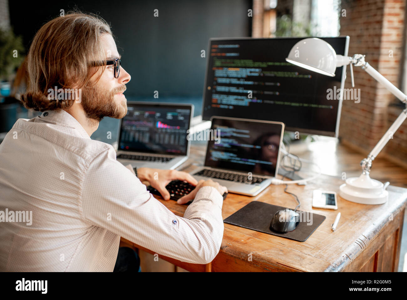 Young male programmer writing a program code sitting at the workplace with three monitors in the office Stock Photo