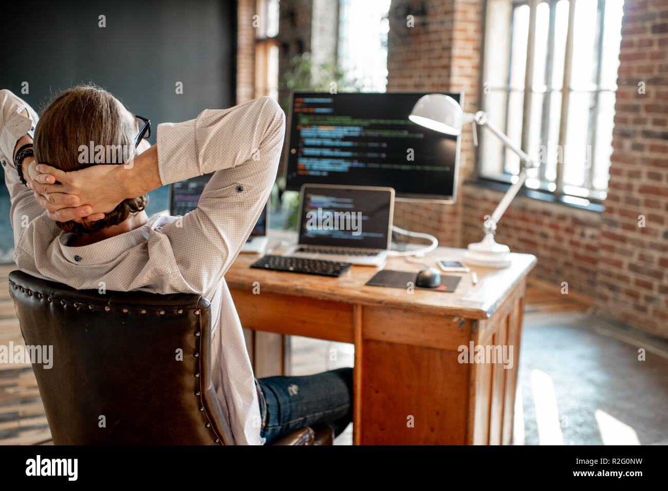 Young male programmer relaxing during the work with program code sitting at the workplace with three monitors in the office Stock Photo