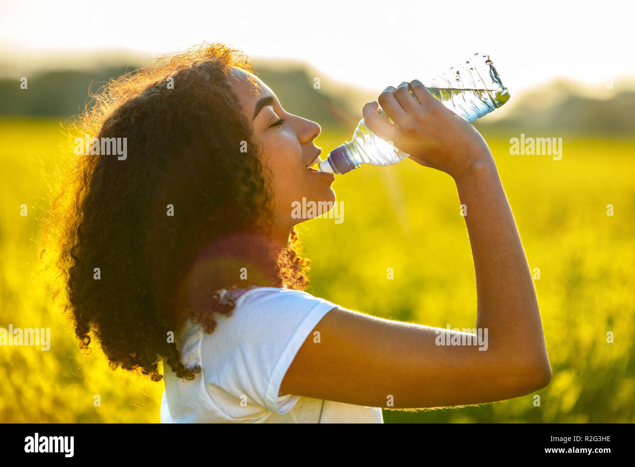 Outdoor portrait of beautiful happy mixed race African American girl teenager female young woman drinking water from a bottle in a field of yellow flo Stock Photo
