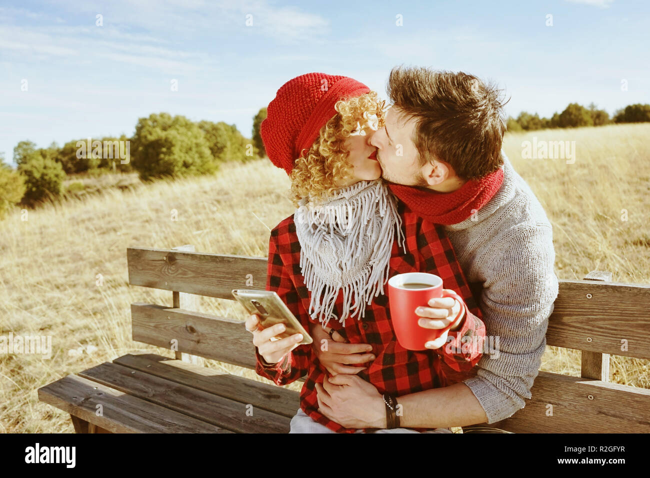 A couple of happy young millennials in love sunbathing on a wooden bench and taking a cup of coffee or tea while they are kissing and holding a smartp Stock Photo