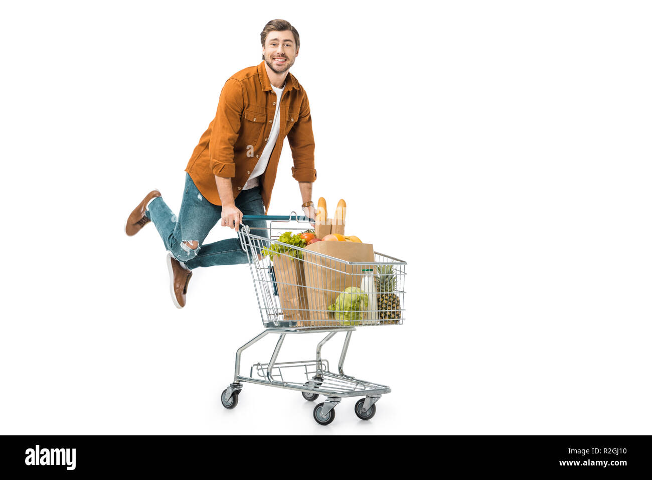 positive man jumping and carrying shopping trolley with products in paper bags isolated on white Stock Photo