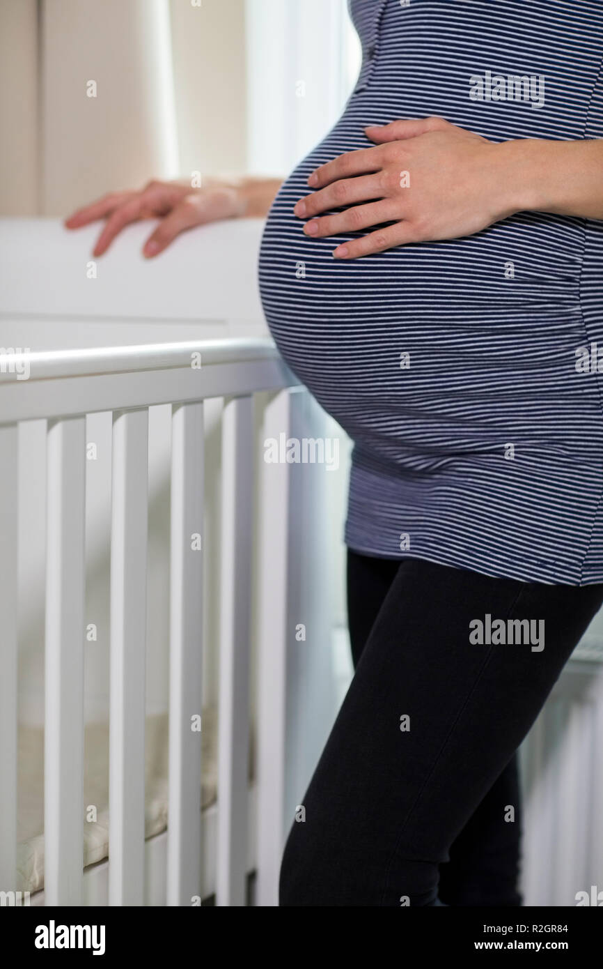 Close Up Of Pregnant Woman Touching Stomach Standing Next To Cot In Nursery Stock Photo