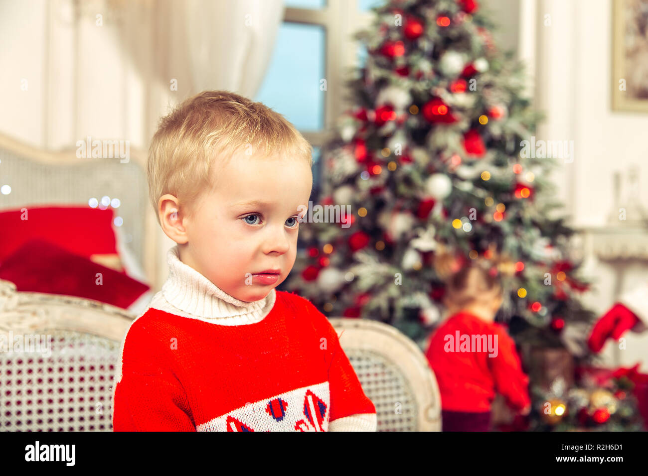 Little sad boy in a festive bedroom on the background of the Christmas tree. Stock Photo