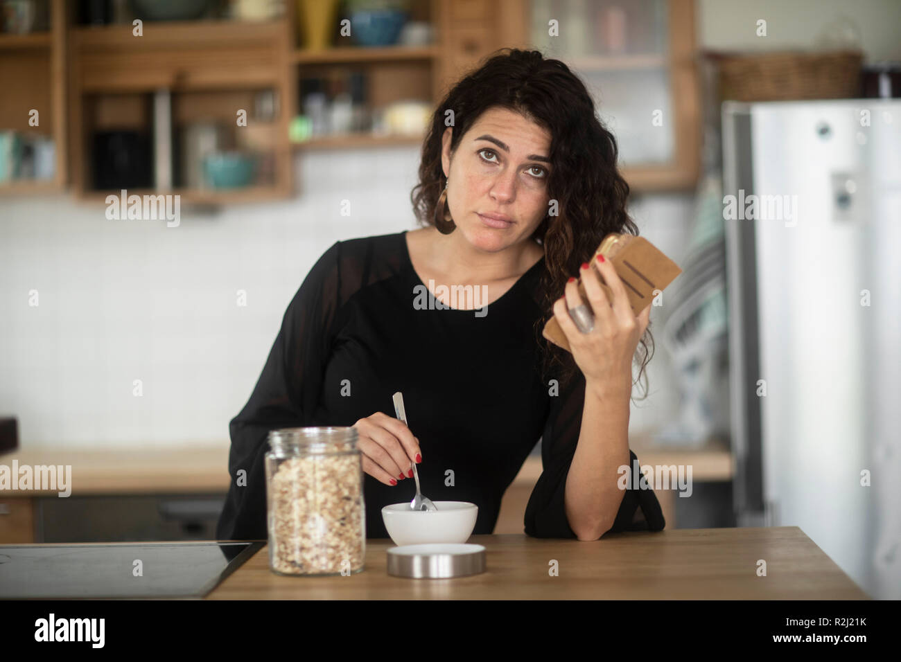 Confused Woman sitting at kitchen counter eating breakfast while looking at her mobile phone Stock Photo