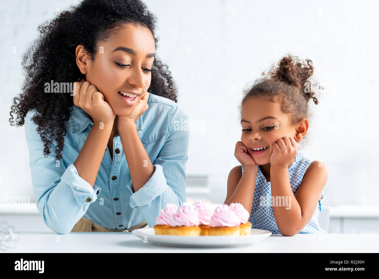 cheerful african american mother and daughter resting chins on hands and looking at cupcakes on table in kitchen Stock Photo