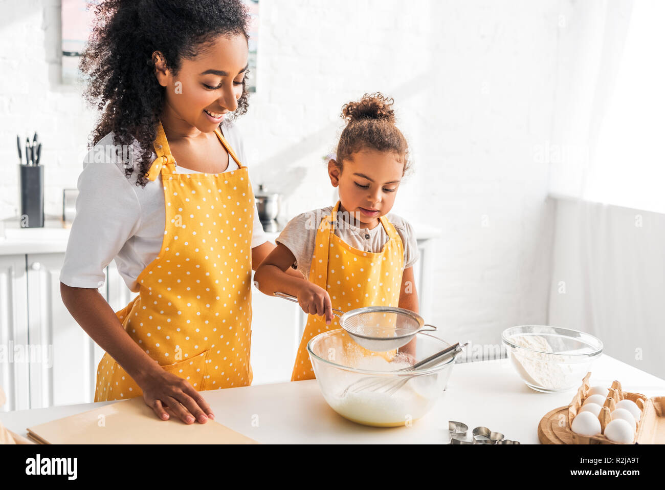 cheerful african american mother and daughter preparing dough and sieving flour in kitchen Stock Photo