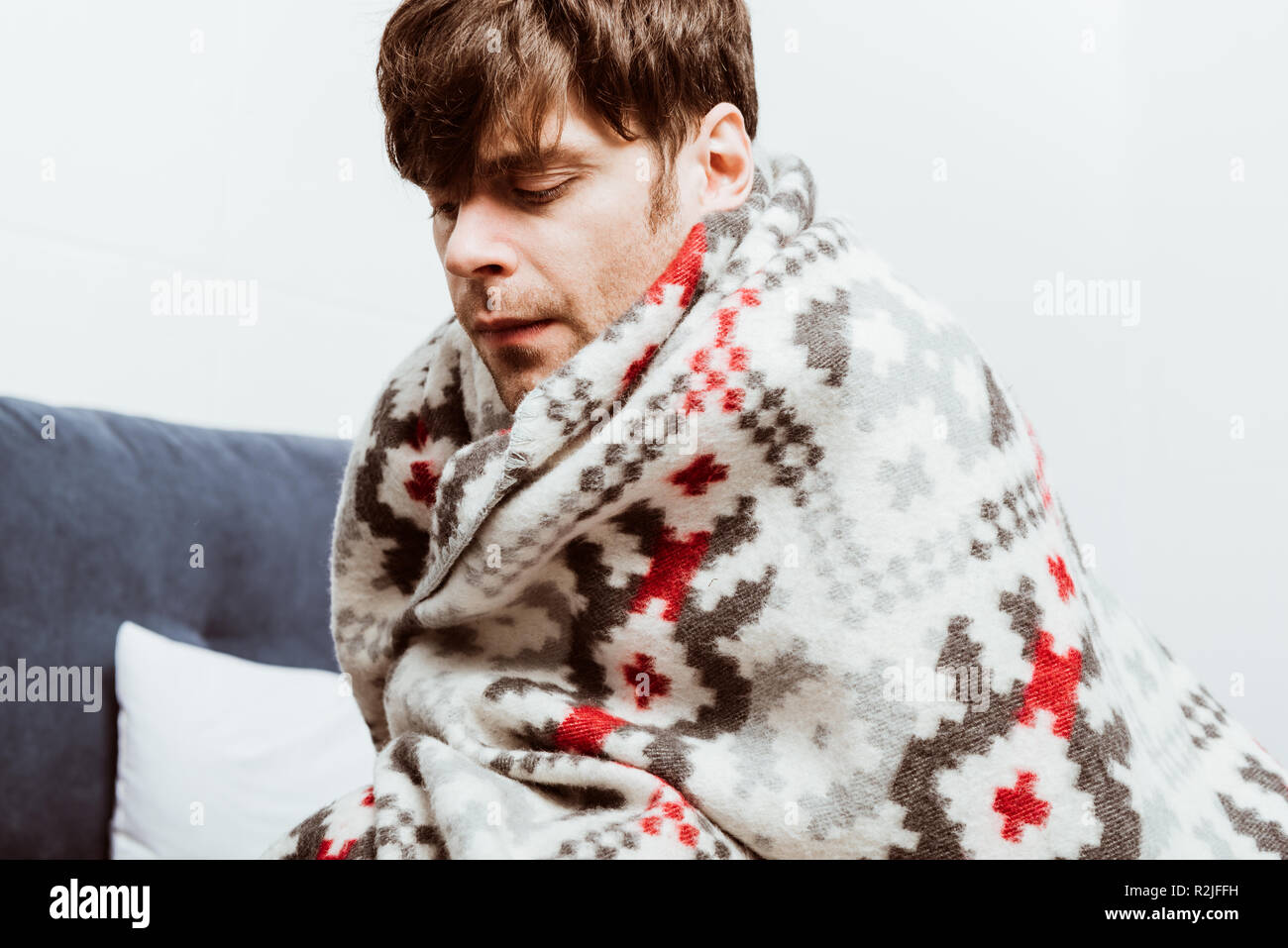 sick young man wrapped in blanket sitting on bed at home Stock Photo