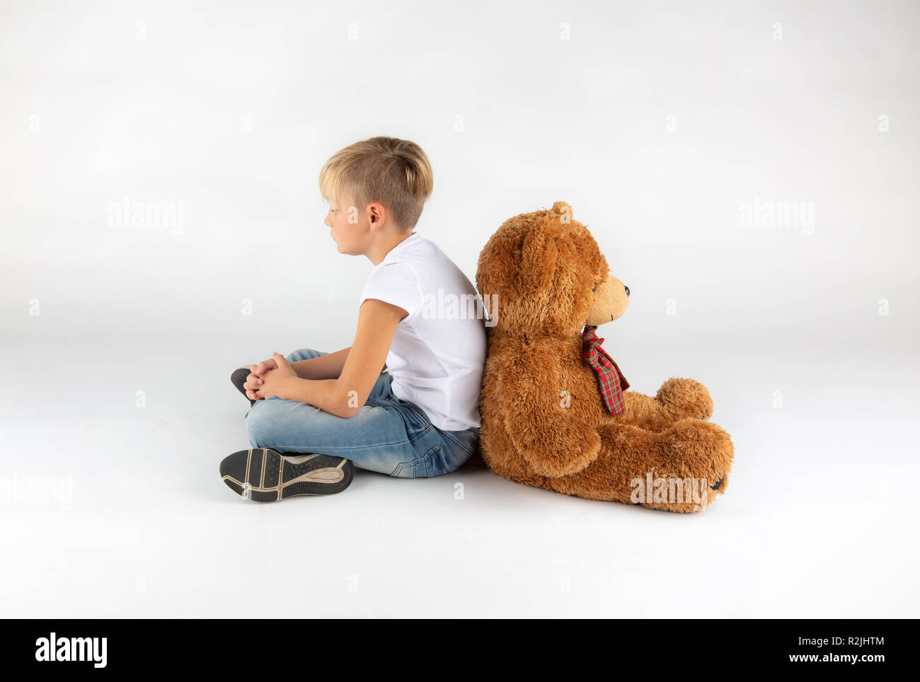 little blond boy is sitting back to back on the floor with a big teddy bear Stock Photo