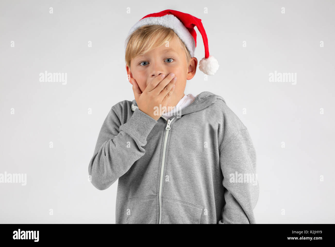 little blond boy with a santa hat is surprised and is covering his mouth with one hand Stock Photo
