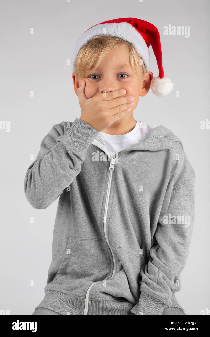 little blond boy with a santa hat is surprised and is covering his mouth with one hand Stock Photo