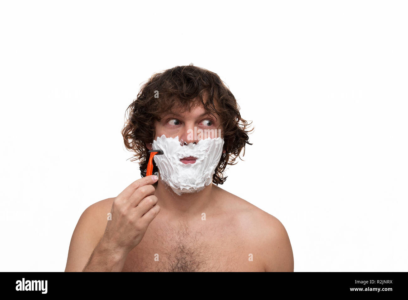 Handsome man with dark wet curly hair shaving his beard with a razor isolated on white. Close up portrait of a man distorts the facewith shaving foam. looking to the right. Copy space. Stock Photo