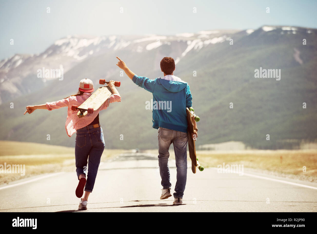 Happy couple is walking by straight road with skateboards or longboards on background of mountains Stock Photo