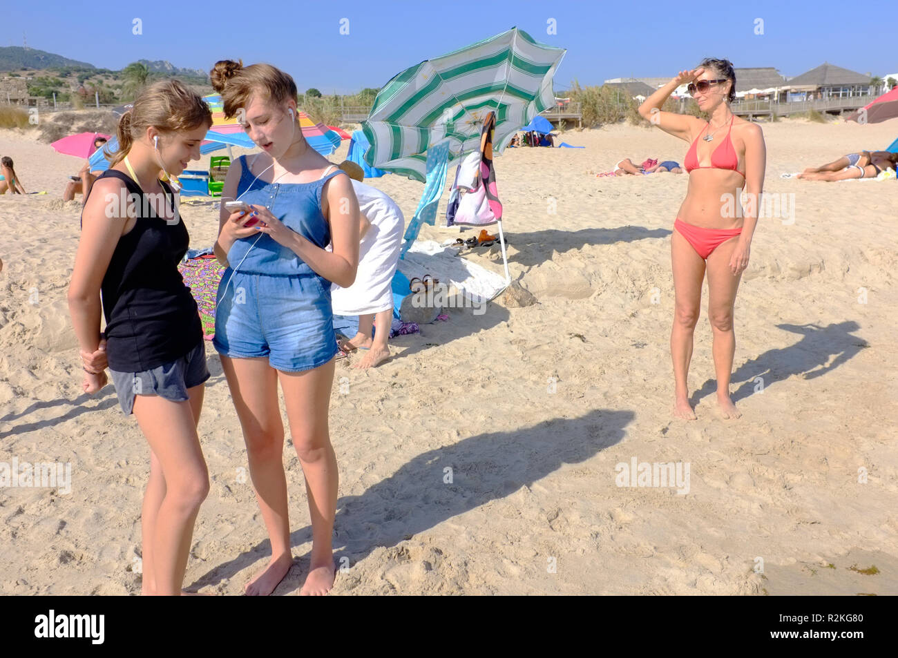 Teenage girls listening to a phone on a beach with their mother in the background. Stock Photo