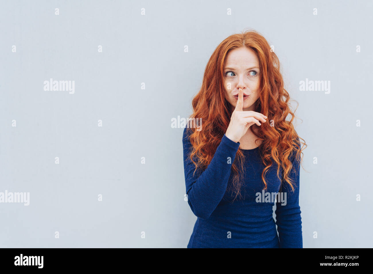 Secretive young woman making a hushing gesture with her finger to her lips as she glances to the side against a white exterior wall with copy space Stock Photo
