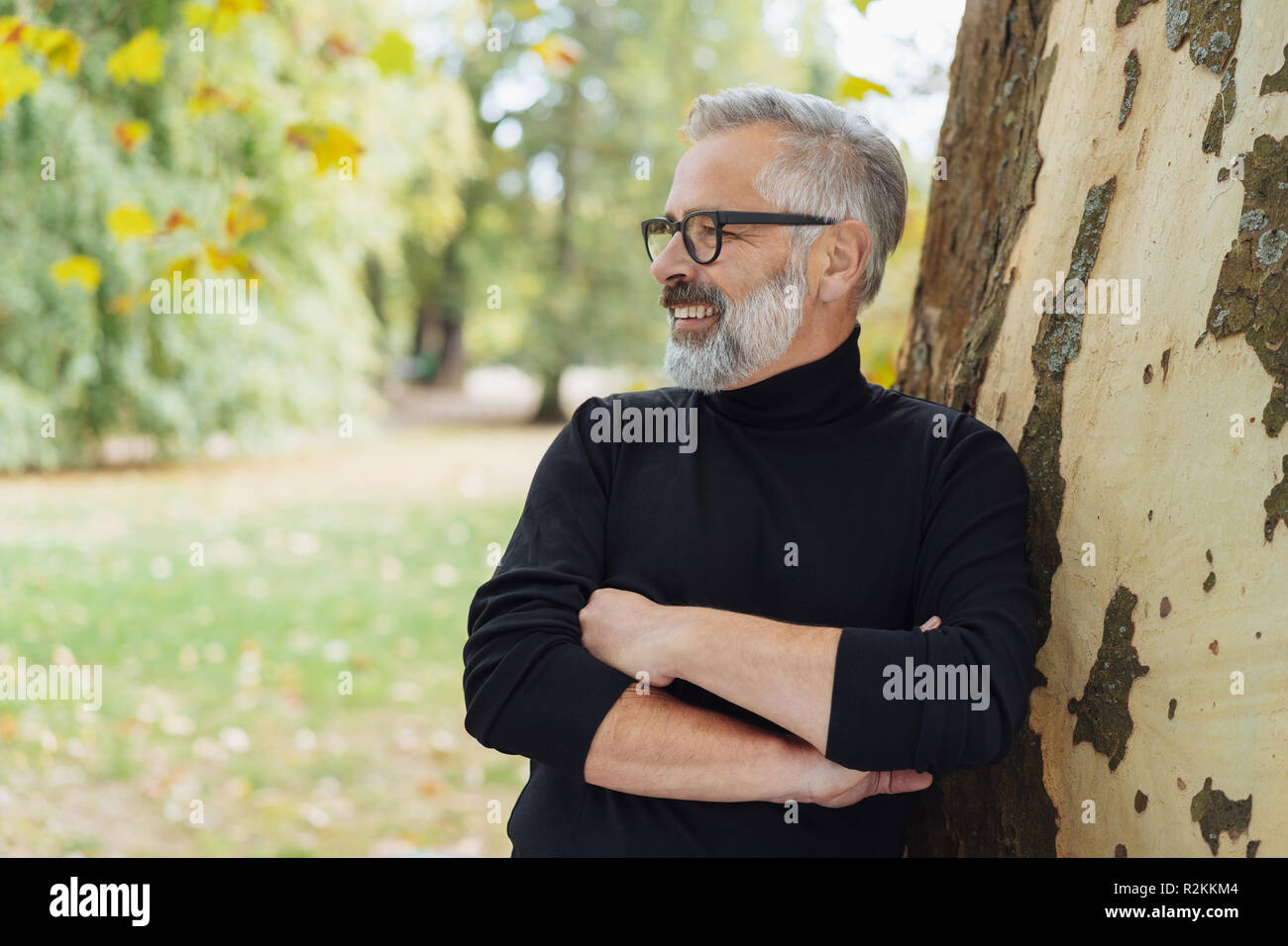 Smiling man with beard and glasses leaning against a tree trunk in an autumn park with folded arms looking to the side Stock Photo