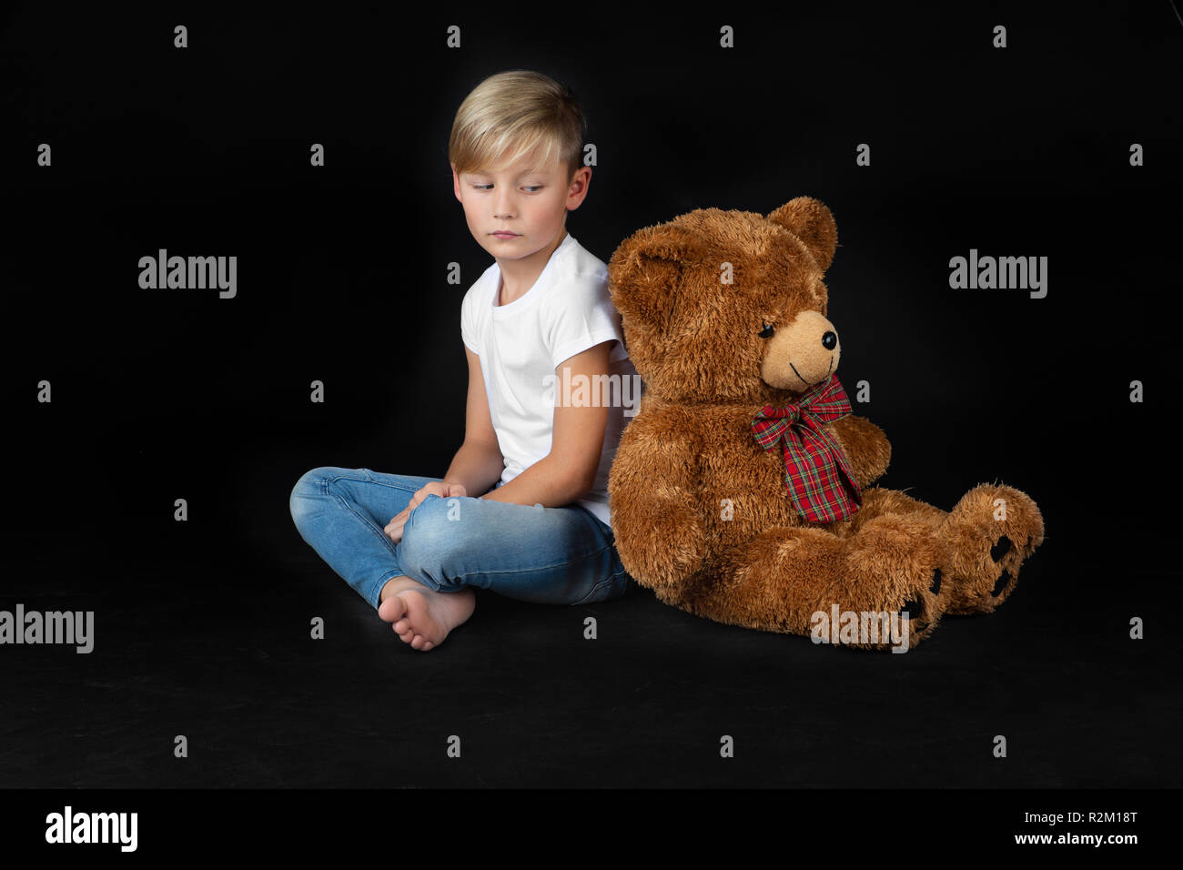 little blond boy is sitting sadly on the floor with a big teddy bear ...