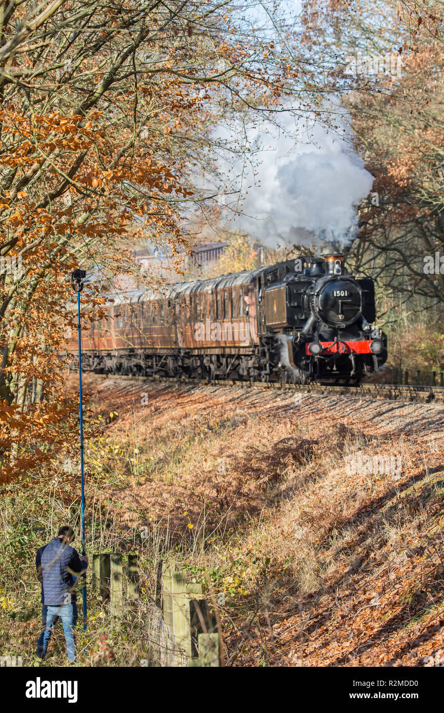 Portrait shot of UK steam train on Severn Valley Railway heritage line, puffing through autumn trees. Man takes elevated photo with camera up on pole. Stock Photo