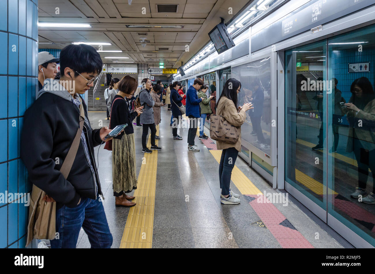 Passengers line up orderly, play on mobile phones and wait for a train on the platform at Samgakji Metro Station in Seoul, South Korea. Stock Photo