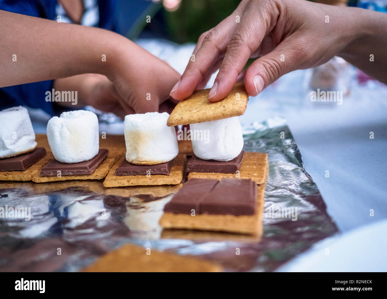An Adult and kids preparing roasted marshmallow cookie sandwich on a picnic table during summertime. Stock Photo