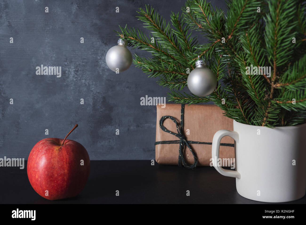 Red apple on a table near a cup with pine branches in it and Xmas balls and a gift box. Christmas frame. Xmas tradition. Winter holidays context. Stock Photo