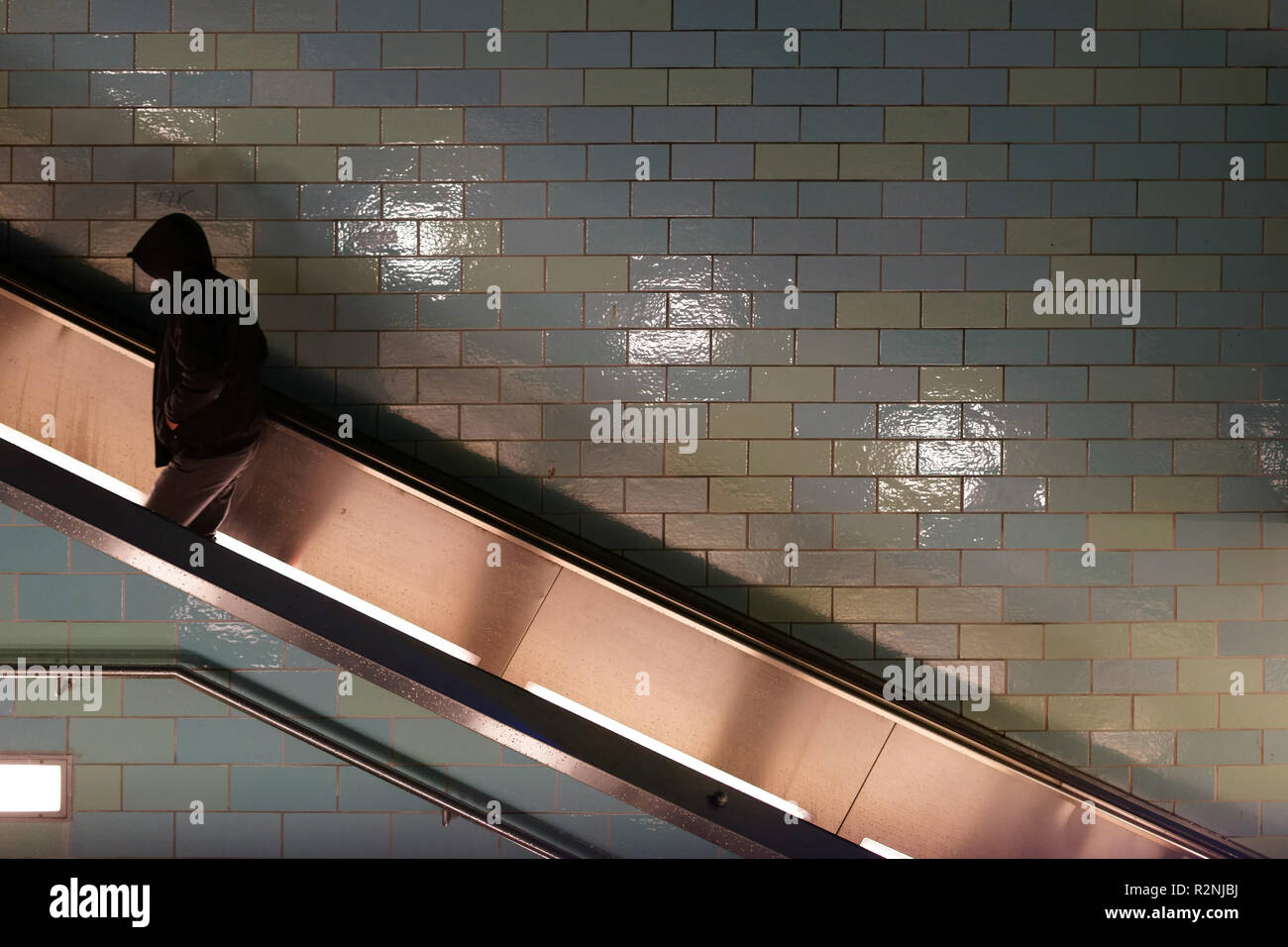 Young man in a hooded jacket goes up a tiled escalator, Stock Photo
