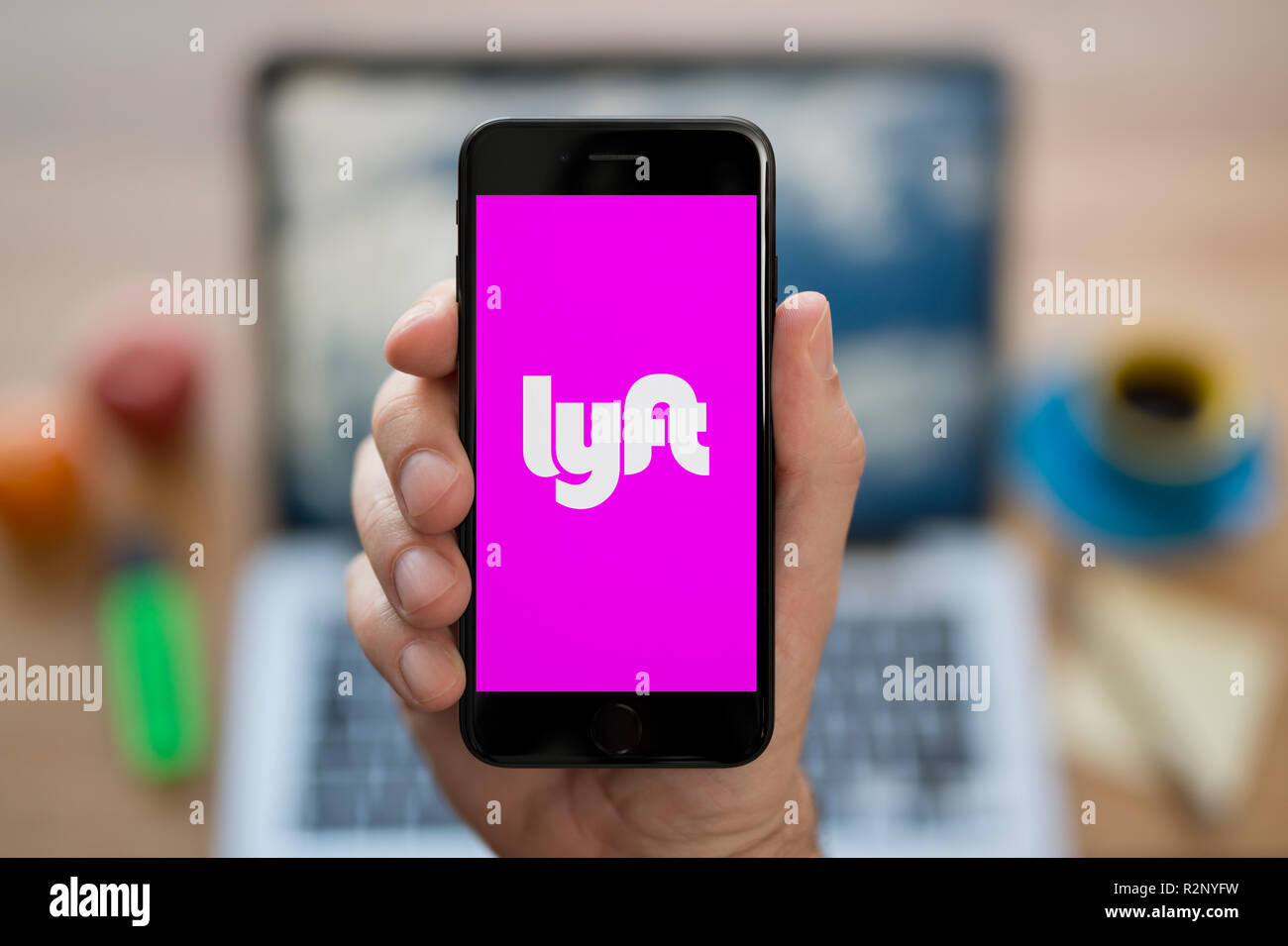 A man looks at his iPhone which displays the Lyft logo, while sat at his computer desk (Editorial use only). Stock Photo