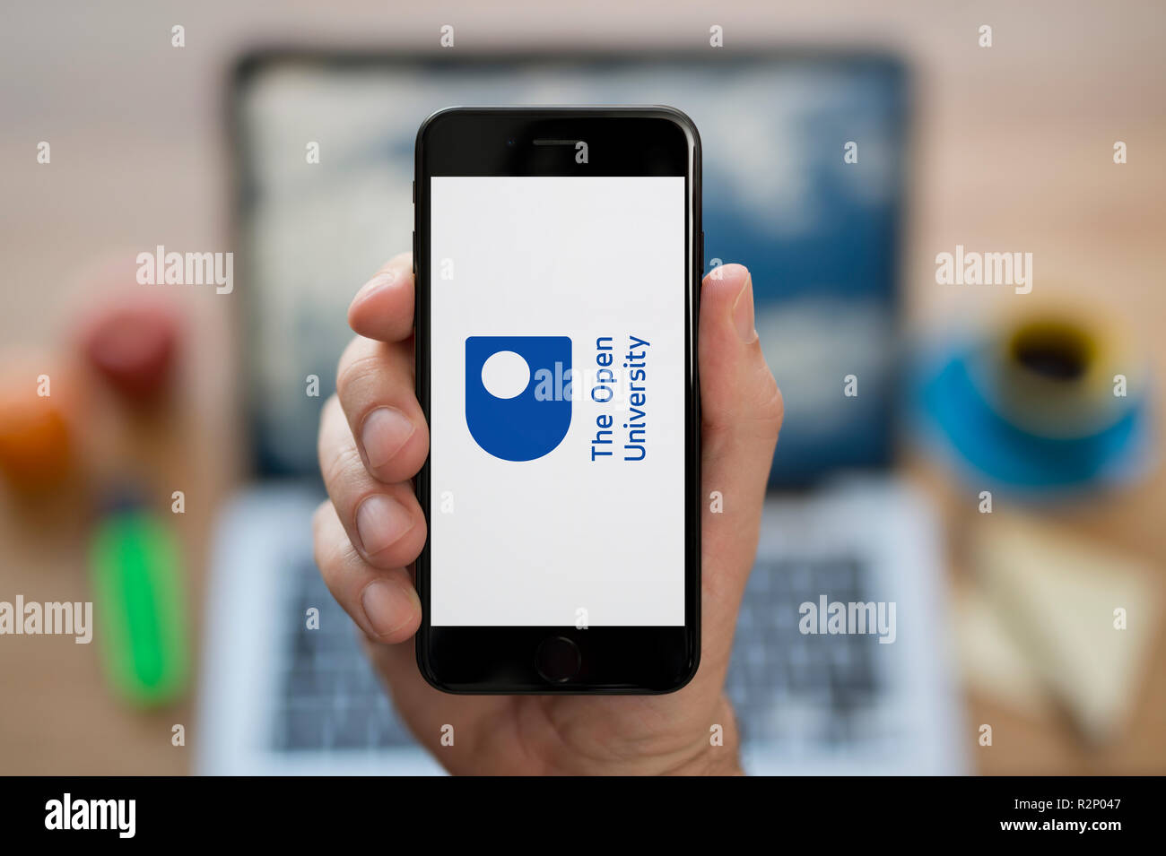 A man looks at his iPhone which displays the Open University logo, while sat at his computer desk (Editorial use only). Stock Photo