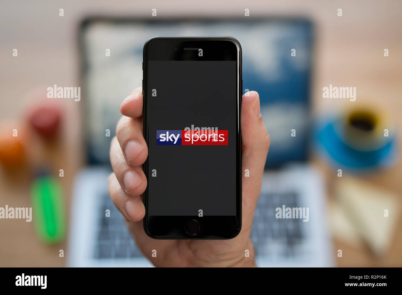 A man looks at his iPhone which displays the Sky Sports logo, while sat at his computer desk (Editorial use only). Stock Photo