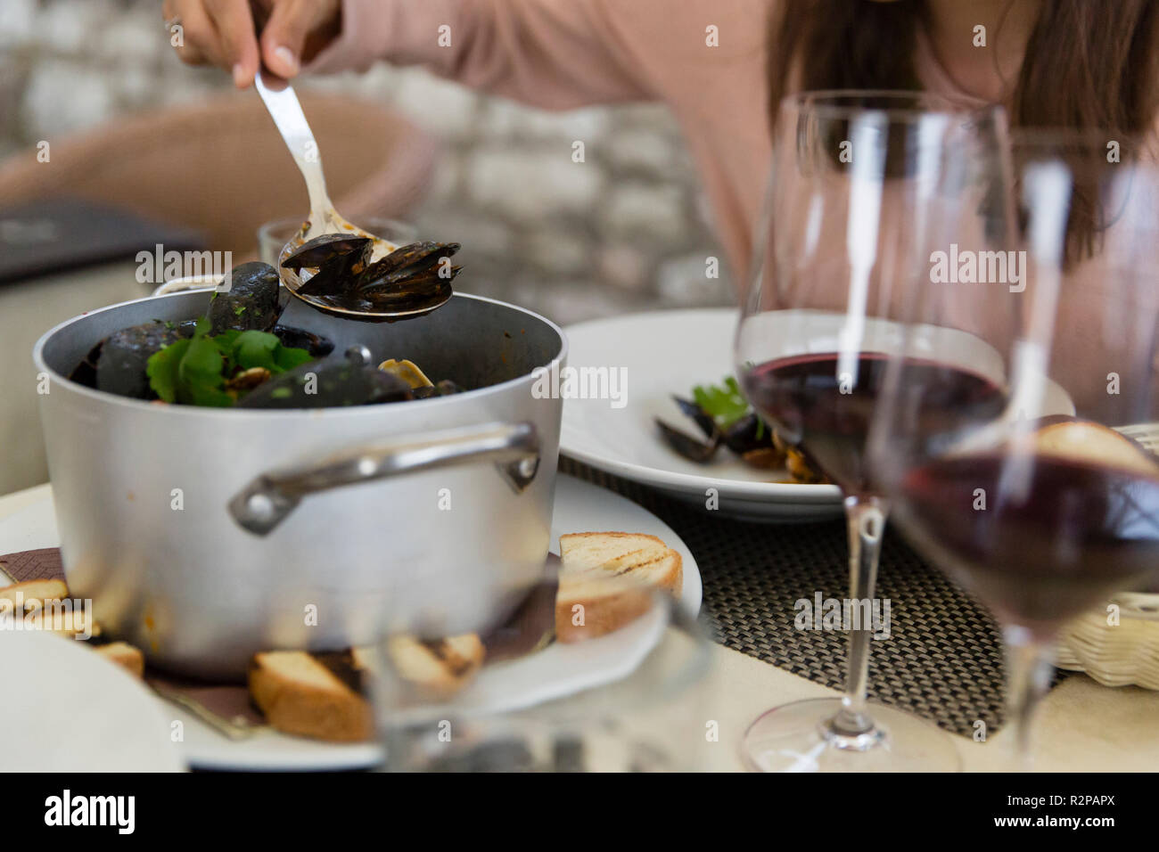 Silver pot full of cooked mussels, woman helps herself, spoon, red wine and white bread on the table Stock Photo