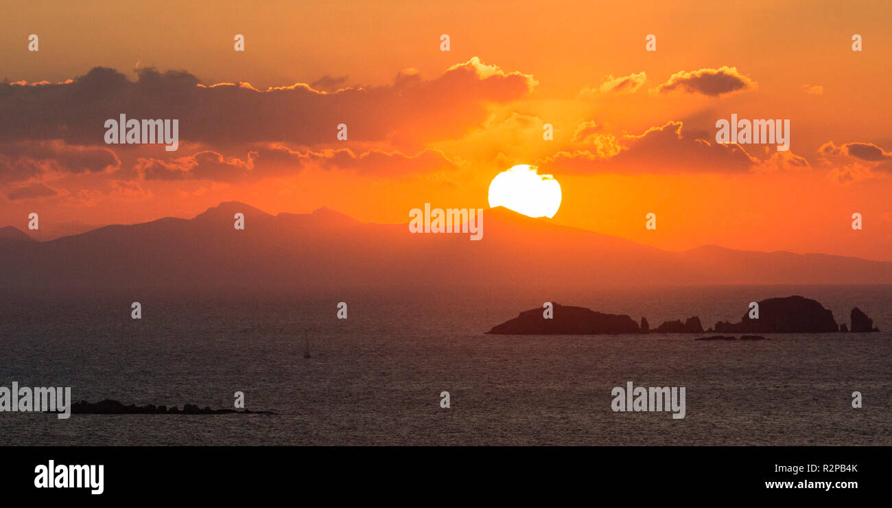Glowing sun above the sea with offshore island and clouds, setting behind a hill, above the sea Stock Photo