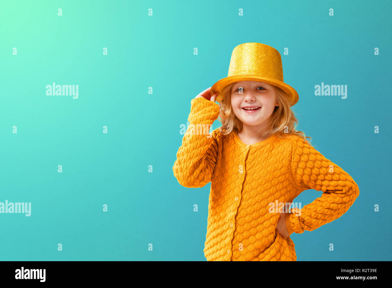 Portrait of a little girl in a knitted mustard jacket and a shiny yellow cylinder hat of a magician on a turquoise background. Holiday concept. Stock Photo