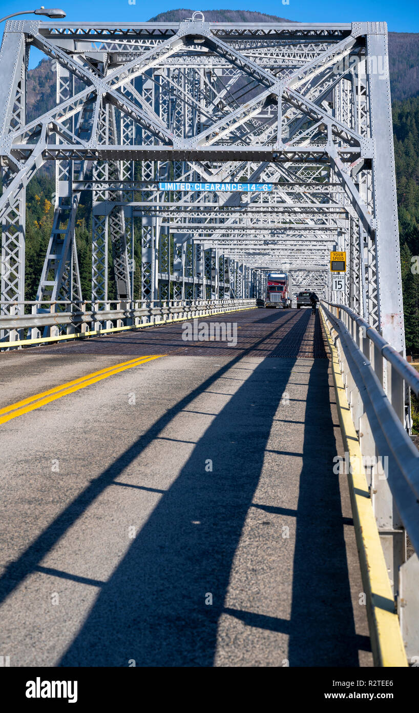 Classic American bonnet big rig burgundy semi truck with shiny refrigerator semi trailer with reefer unit on it driving trough truss Bridge of God in  Stock Photo