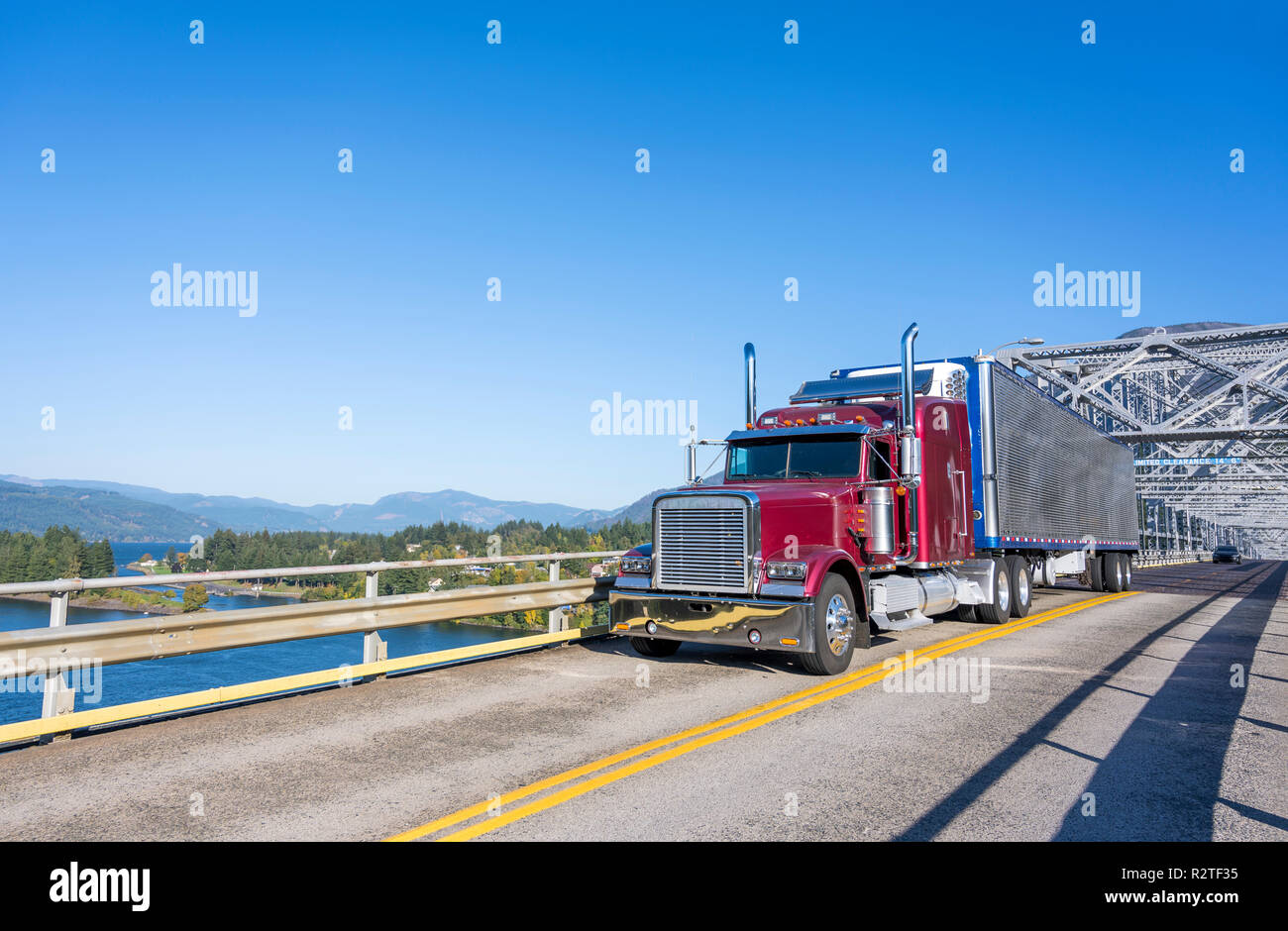 Classic American big rig burgundy semi truck with refrigerated semi trailer with reefer unit on it driving on truss Bridge of God with view of Columbi Stock Photo