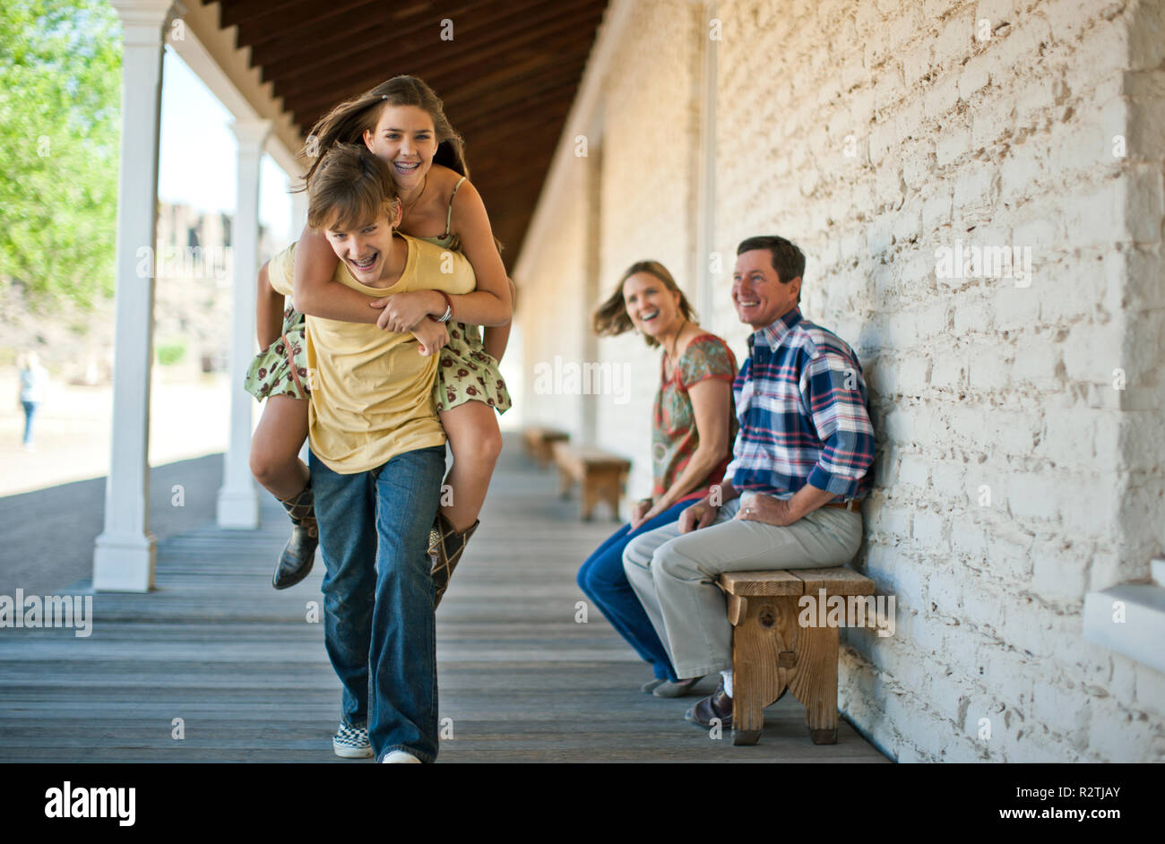 Smiling teenage boy  carrying his sister on his back as their parents look on. Stock Photo